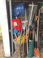 Overall view of garbage can full of long-handled tools, snow shovels, rakes, extension cord visible