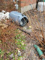 Gray plastic rain barrel on ground with lid, spigot visible, overflow hose nearby