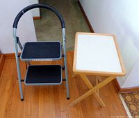 Steel two-step folding step stool beside light-wood folding snack tray table
