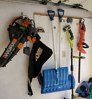 Overall lot hanging on garage wall: WORX WG500 blower with bag, Ryobi trimmer, Black & Decker hedge trimmer, two blue snow shovels, battery and charger visible
