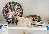 Shelf view showing Hummel plate on stand with certificate and stack of other plates