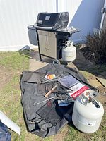 Grill with attached tank, spare tank, cover, tools and baskets on ground