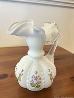 Full view of milk glass pitcher showing floral design, ribbing, clear handle and ruffled rim