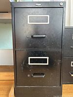 Front view of black metal two-drawer filing cabinet showing handles, label frames and lock cylinder