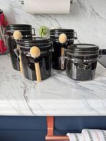Front view of four black ceramic canisters with three wood spoons attached