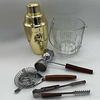 Group shot of all items: gold shaker, glass ice bucket, strainer, jigger, tongs