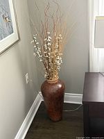 Full view of brown floor vase with dried stem arrangement beside wall and furniture