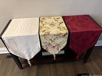 Three polyester table runners in white/ivory, gold/beige floral, red/burgundy paisley displayed on console table