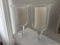 Both clear glass hurricane candle holders with included white pillar candles displayed side by side