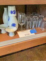 Cabinet shelf containing porcelain sake bottles, stem wine glass, upside-down clear glass tumblers and porcelain pedestal cups