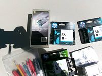 Lot overview showing battery box, HP 56 cartridges, HP 65XL cartridge, Epson cartridge foil pack, bag of markers and pens