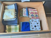 Drawer view of folded linens including packaged napkins, striped cloths, plaid napkins, patchwork pieces, cat-print napkins