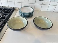 Stack of six teal-rimmed plates beside two speckled interior soup bowls