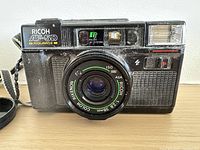 Front view of Ricoh AF-5D camera showing lens, flash, branding, attached strap and lens cap