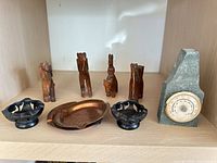 Group view of four wooden animal figurines, two black soapstone dishes, copper ashtray, and stone thermometer on shelf