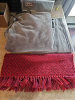 Grey fleece blanket stacked with red woven rug showing fringe and pattern