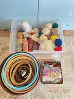 Plastic tote with yarn balls and floss tin beside plaid tin of hoops