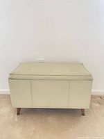 Front view of cream vinyl storage bench showing button-tufted lid and tapered legs