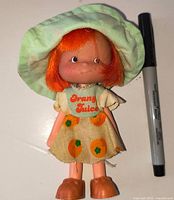 Front view of Orange Juice doll with hat, dress, shoes and scale pen