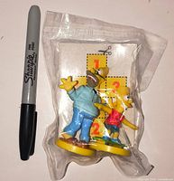 Sealed bag showing Homer and Bart Simpson promo figures beside a Sharpie for scale