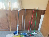 Full view of eight long-handled cleaning tools leaning against wall