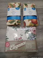 Two packaged Mainstays tablecloths and one packaged Elegance queen sheet set on floor