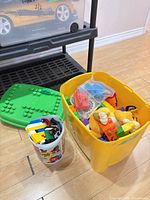 Yellow storage bin containing large multi-colour blocks beside green base plate and clear bucket of small blocks, Simba plush visible