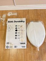 Kohler box beside wrapped elongated toilet seat showing brand and packaging