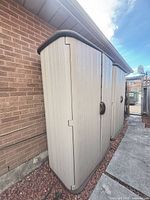 Full exterior view of beige vertical resin shed beside brick wall