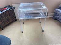 Full view of clear acrylic side table showing top, shelf, and caster wheels