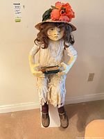 Front view of seated papier-mâché doll on pedestal showing overall appearance, dress, shoes, hat, book