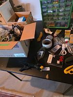 Overview of table with box of wiring and tools, spring clamp, yellow hand tool, tape, abrasive disc card