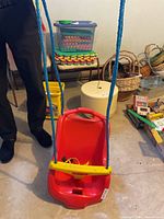 Front view of red Fisher-Price infant swing seat showing yellow bar, safety strap and blue hanging ropes
