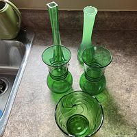 Five green glass pieces on countertop: two matching flared vases, two individual tall vases, one small bowl