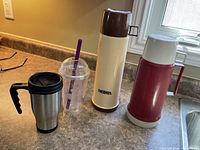 Four beverage containers on countertop: stainless travel mug, clear tumbler, beige Thermos bottle, red insulated bottle