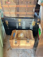 Three stacked trunks: brown canvas-covered on top, black canvas-covered in middle, natural wood crate trunk at bottom