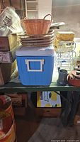 Blue cooler with nested wicker baskets on top, rack and small items adjacent