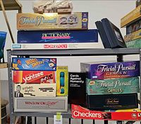 Shelf with multiple boxed games including Trivial Pursuit, Pictionary, Monopoly, Cards Against Humanity, Trivial Pursuit Genus V, Battleship cases, Jenga blocks
