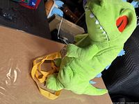 Front view of green Reptar plush backpack standing upright on box showing orange straps and facial details