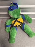 Full view of large Leonardo TMNT plush sitting upright, showing green body, yellow chest, blue mask and harness straps