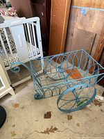 Side view of blue wrought iron decorative wheelbarrow planter showing wheels, basket, and handle