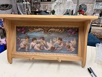 Front view of wood wall shelf showing cherub wallpaper inset and two pegs