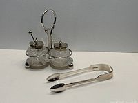 Sterling tongs beside silver-plated condiment stand with two glass jars and spoon