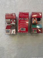 Three clear plastic packages each containing multiple flat-packed miniature houses, back panels show product text for 24 miniature houses