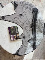 Unfolded black mesh spider web mantel scarf alongside its packaging