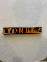 Front view of orange wooden sign with black text THE WITCH IS IN THE HOUSE