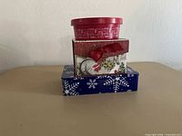 Three stacked holiday-themed treat containers: red plastic tub on white cardboard gift box on blue metal tin