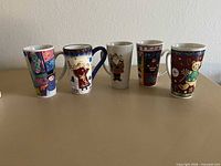 Five tall Christmas-themed ceramic mugs lined up to show different designs and height.
