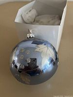 Metallic blue glass ornament with silver leaf and glitter dot design next to white box