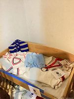 Overview of assorted baby clothing in crib including blue striped sweater, white cardigan, light blue sweater